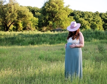 Pregnant woman with tan smock holding belly surrounded by green bushes.