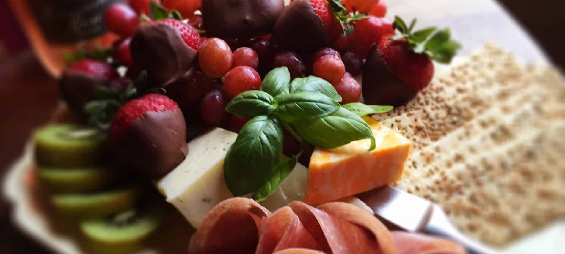 White tray piled with chocolate covered strawberries, white and orange cheeses, salami, crackers, and kiwi