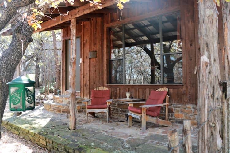 Exterior of rustic cabin with covered porch and sitting chairs, surrounded by trees