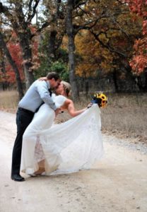 Groom dipping his bride into an embraced kiss on a dir path surrounded by colorful trees in the fall