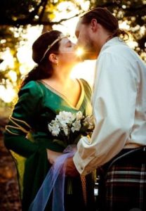 Groom and bride dressed in Scottish attire, kissing at sunset surrounded by trees