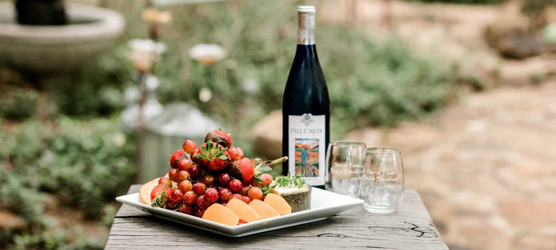 Outdoor table topped with white tray piled with fresh fruit and cheese, bottle of wine, two wine glasses